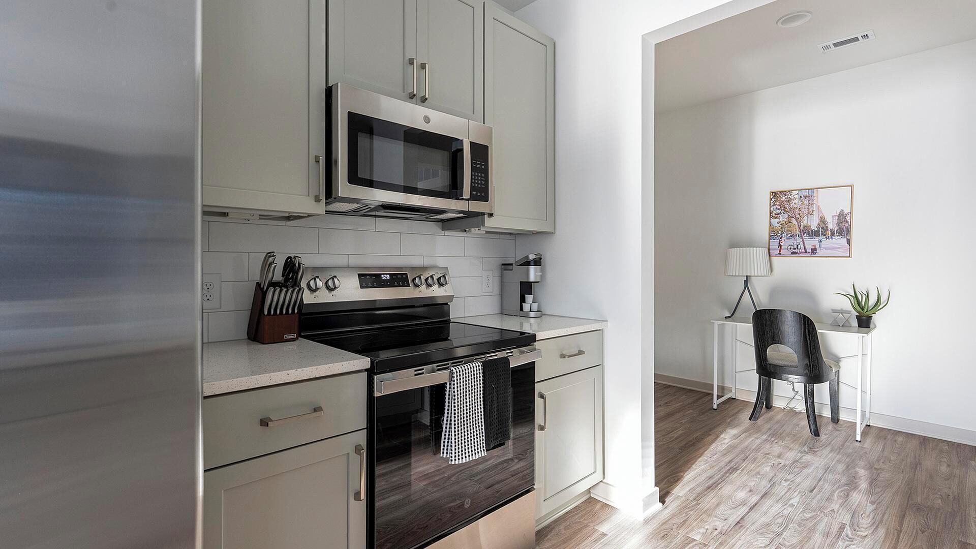 A modern kitchen with a built-in microwave, oven, and coffee maker, a small dining area with a chair and table, and a framed picture on the wall.