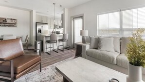 Een moderne woonkamer met een leren bank, een witte salontafel en een grijze bank met kussens. Er is een keukenhoek met een koelkast, magnetron en barstoelen. In de achtergrond staat een eettafel met stoelen.