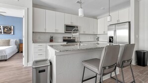 A modern kitchen with white cabinets, a granite countertop, stainless steel appliances, and a bar area with two chairs. - Landing | Modern 2BD, Gym (Fort Myers)