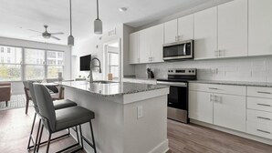 A modern kitchen with white cabinets, a central island, stainless steel appliances, and a dining area with bar stools. - Landing | Modern 2BD, Gym (Fort Myers)