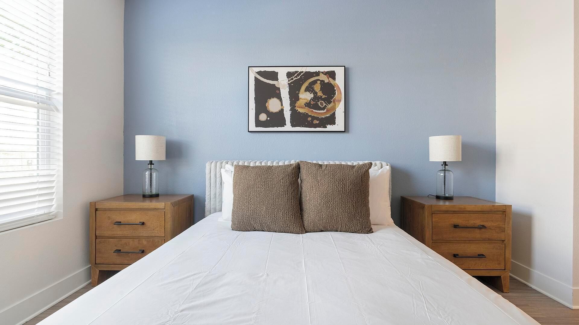 Une chambre à coucher comprenant un lit, deux tables de chevet, une lampe sur chaque table de chevet, une œuvre d’art murale et une fenêtre avec des stores.