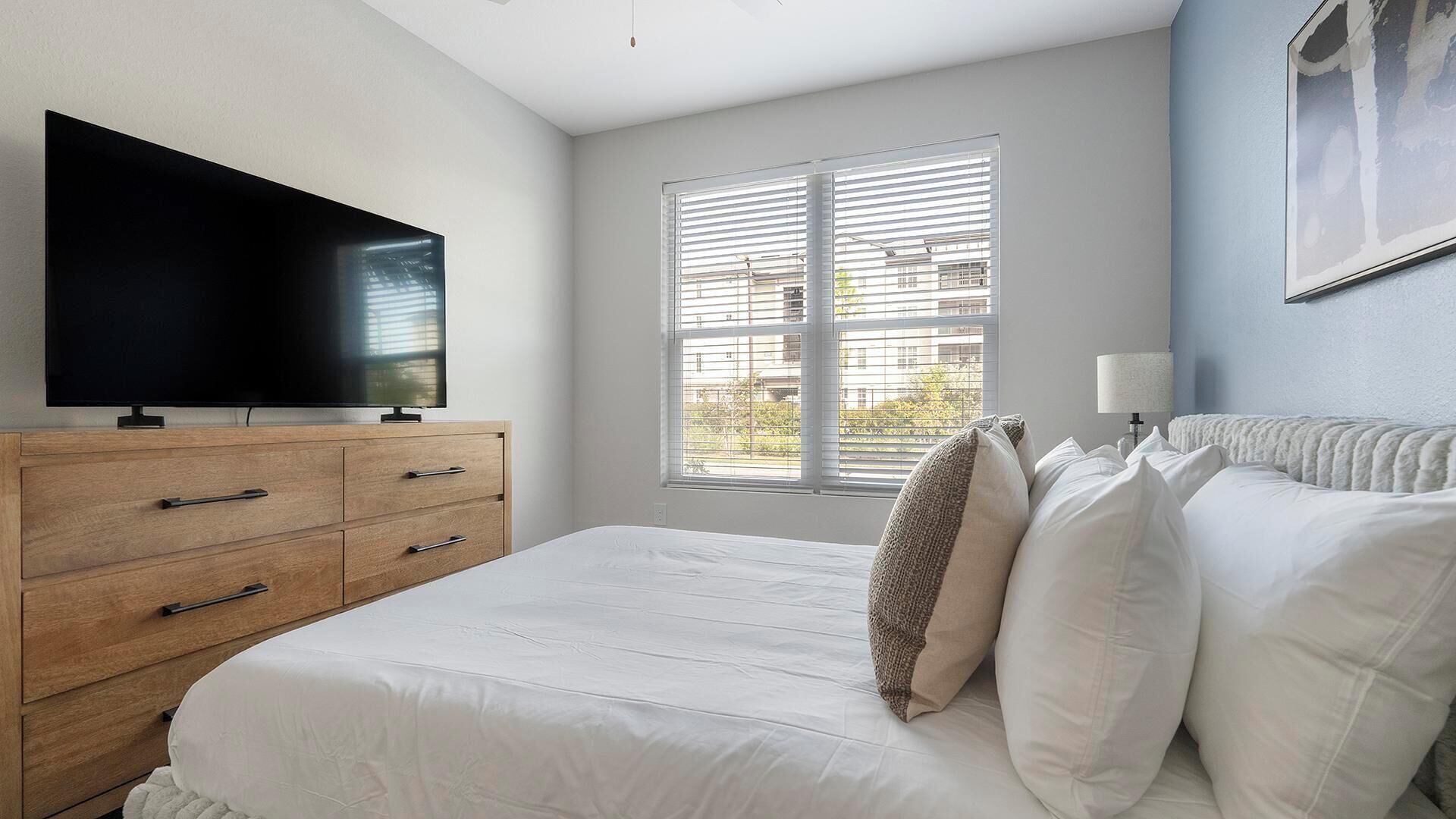 A bedroom with a bed, a TV on a wooden dresser, a window with blinds, and a lamp on the nightstand.