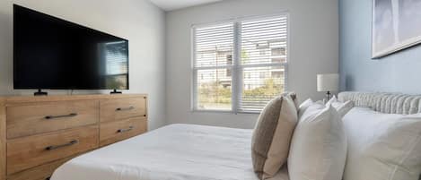 A bedroom with a bed, a TV on a wooden dresser, a window with blinds, and a lamp on the nightstand.
