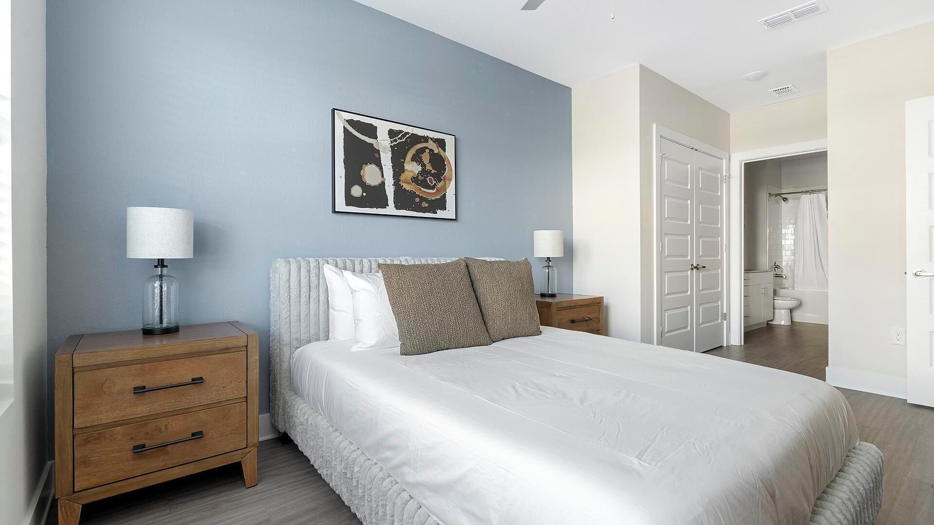 A bedroom with a bed, two bedside tables, a nightstand, a framed picture on the wall, and a bathroom visible through an open door.