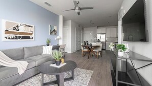 A modern living room with a grey sofa, a coffee table, and a dining area with a wooden table and chairs. There is a kitchen visible in the background with white cabinets and a large TV mounted on the wall. - Landing | Beautiful 2BD, Gym (Fort Myers)