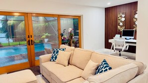 Uma sala de estar com um sofá bege, uma escrivaninha com um computador e vista para uma piscina através de portas de vidro.