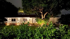 Uma casa com uma árvore grande na frente, cercada por arbustos e grama bem cuidados.