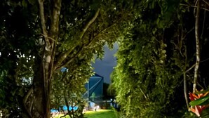 Uma trilha externa iluminada à noite, ladeada por árvores e vegetação, conduz a um edifício com uma estrutura azul ao fundo.