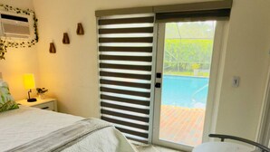 Quarto com cama, mesa de cabeceira com abajur, porta de vidro que dá para uma área de piscina e uma pequena mesa redonda com cadeiras.