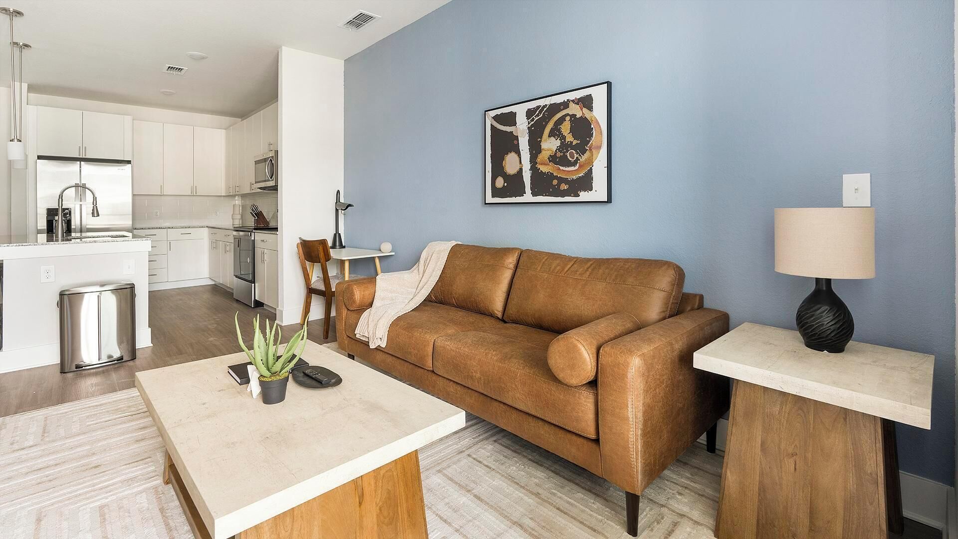 Una sala de estar moderna con un sofá de cuero marrón, una mesa de centro, una lámpara y una cocina al fondo.