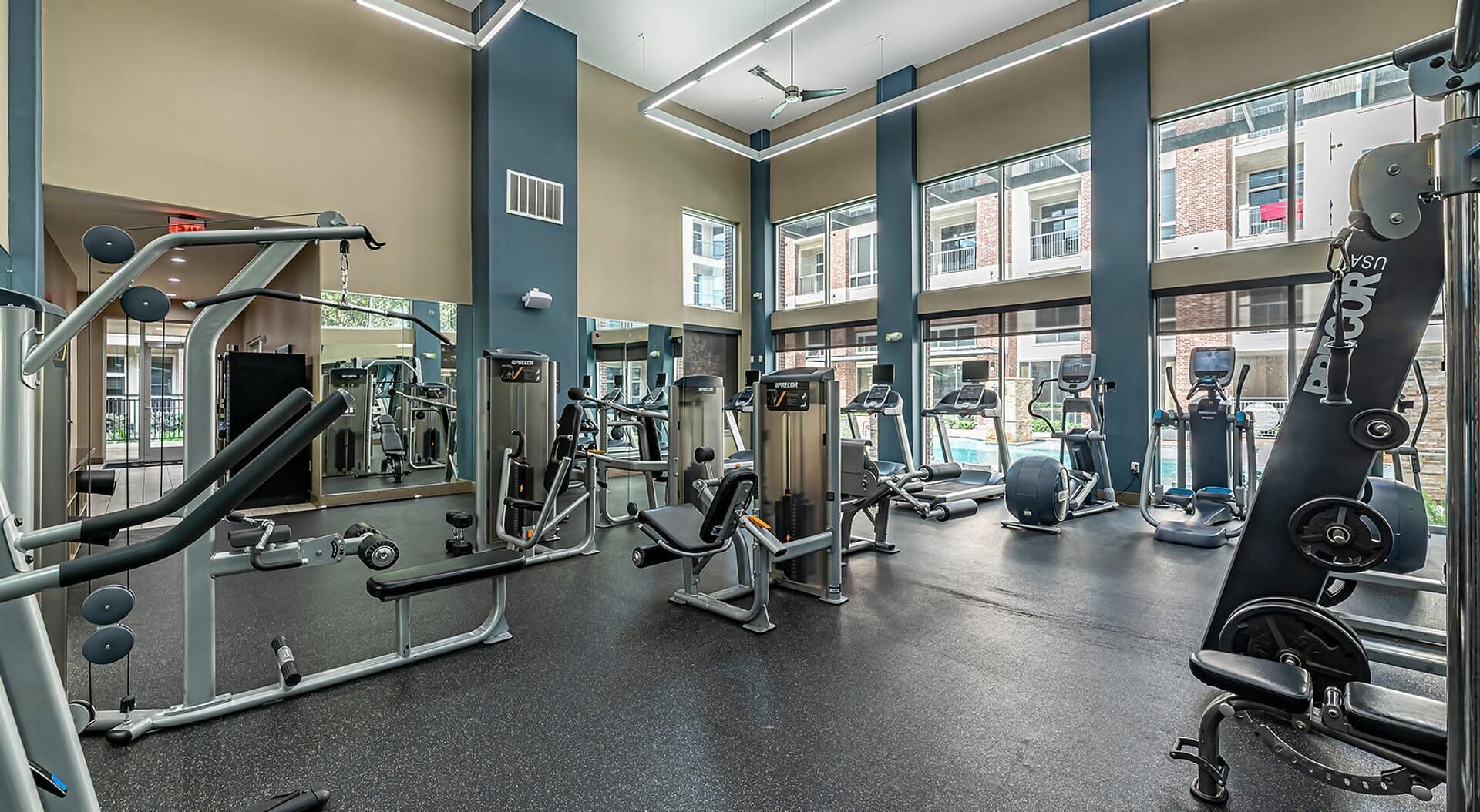 A gym with various exercise machines, large windows, and a high ceiling.