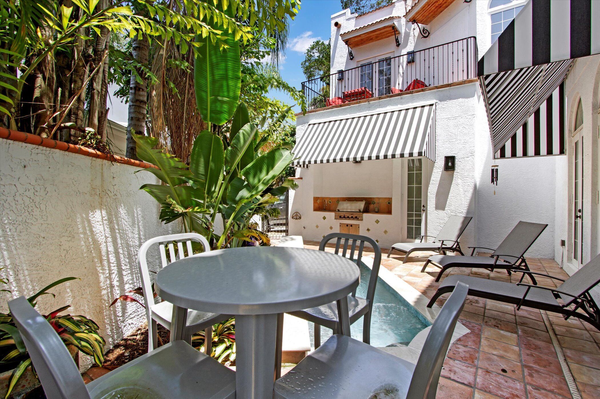 A poolside area with a round table and chairs, a barbecue grill, and a view of a building with striped awnings.