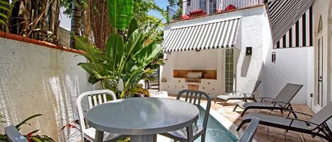 A poolside area with a round table and chairs, a barbecue grill, and a view of a building with striped awnings.