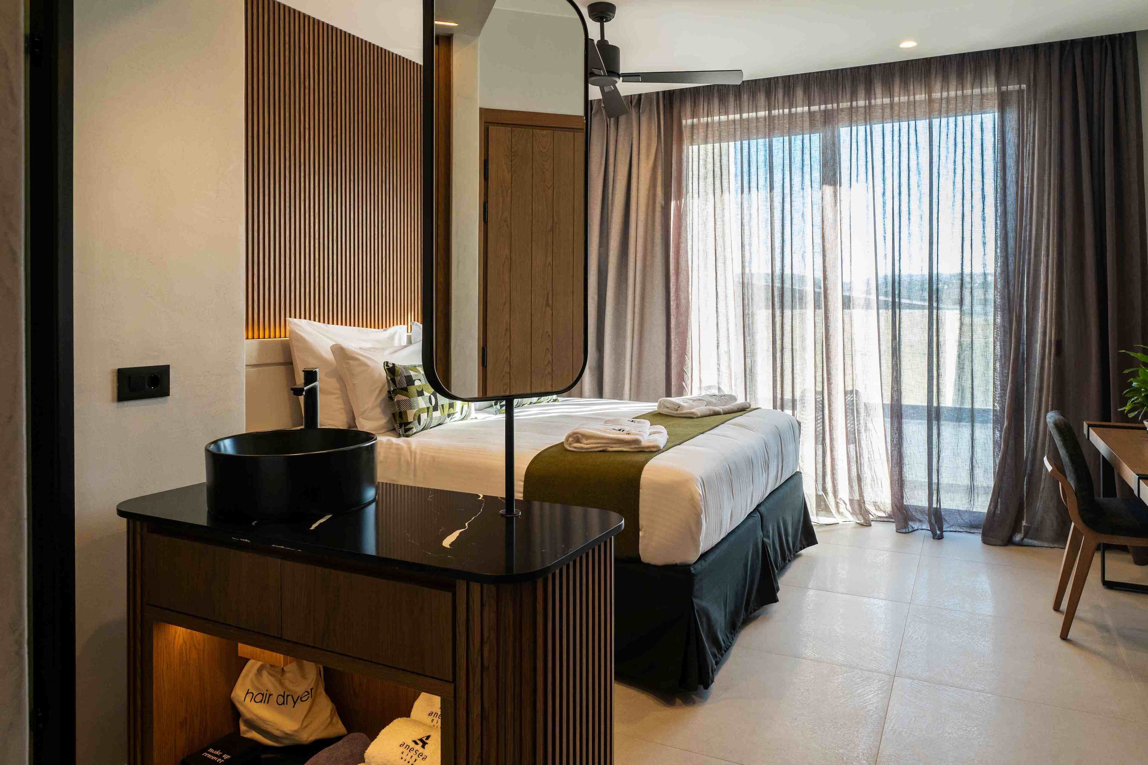 Habitación de hotel con cama, escritorio con espejo, secador de pelo y vistas al exterior a través de amplias ventanas.