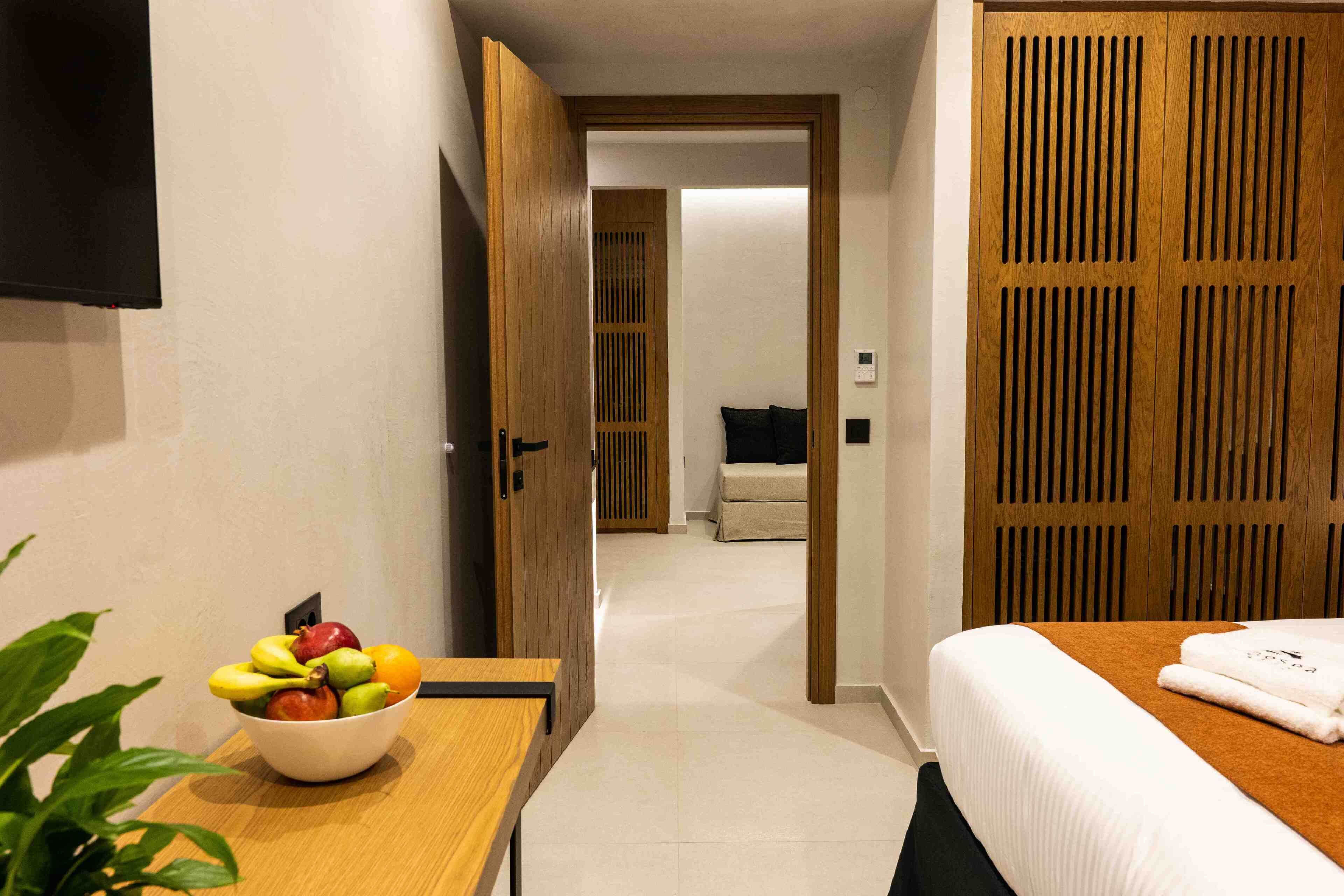 Habitación de hotel con una cama, una mesita de noche con un frutero, un armario de madera y un televisor en la pared.