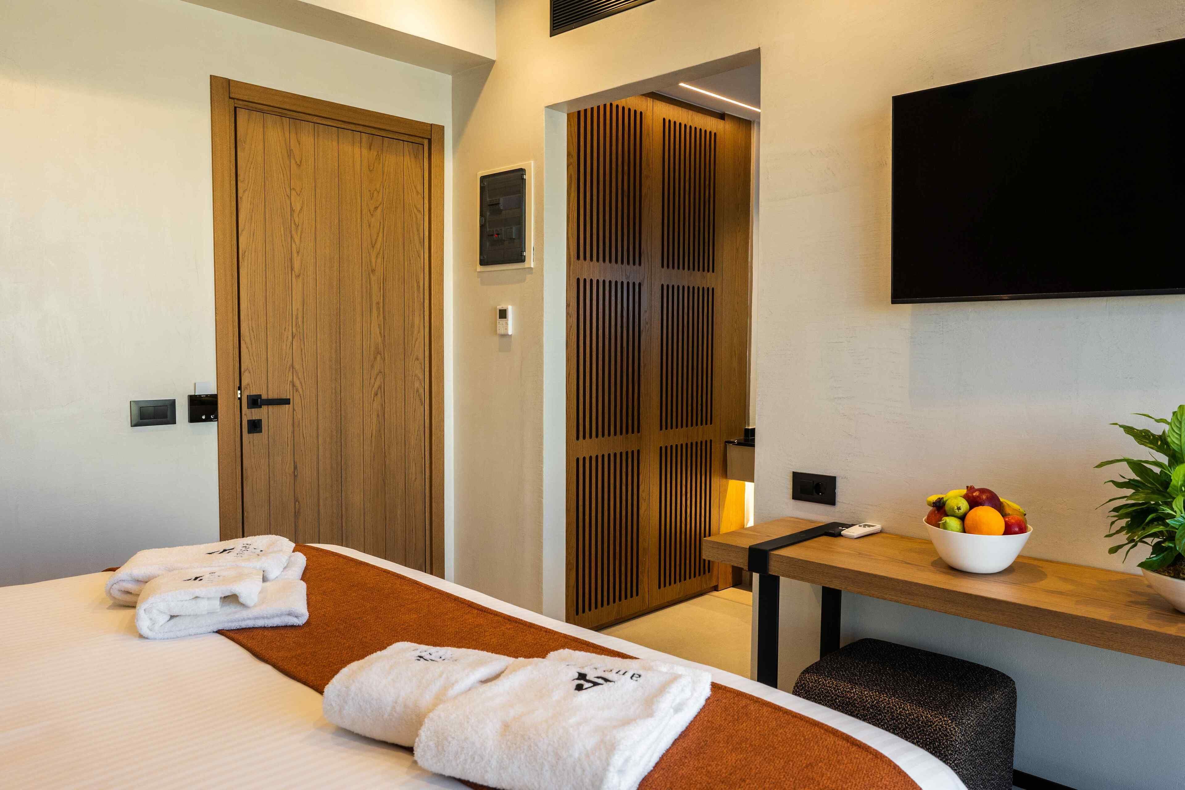 Een hotelkamer met een bed, een houten garderobe, een aan de muur bevestigde televisie, een tafeltje met een schaal fruit, en een potplant.