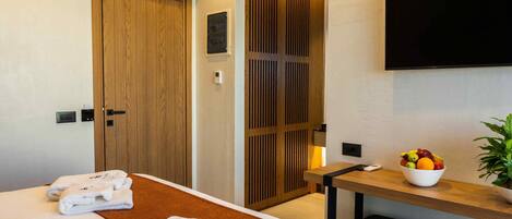 A hotel room with a bed, a wooden wardrobe, a TV mounted on the wall, a small table with a fruit bowl, and a potted plant.