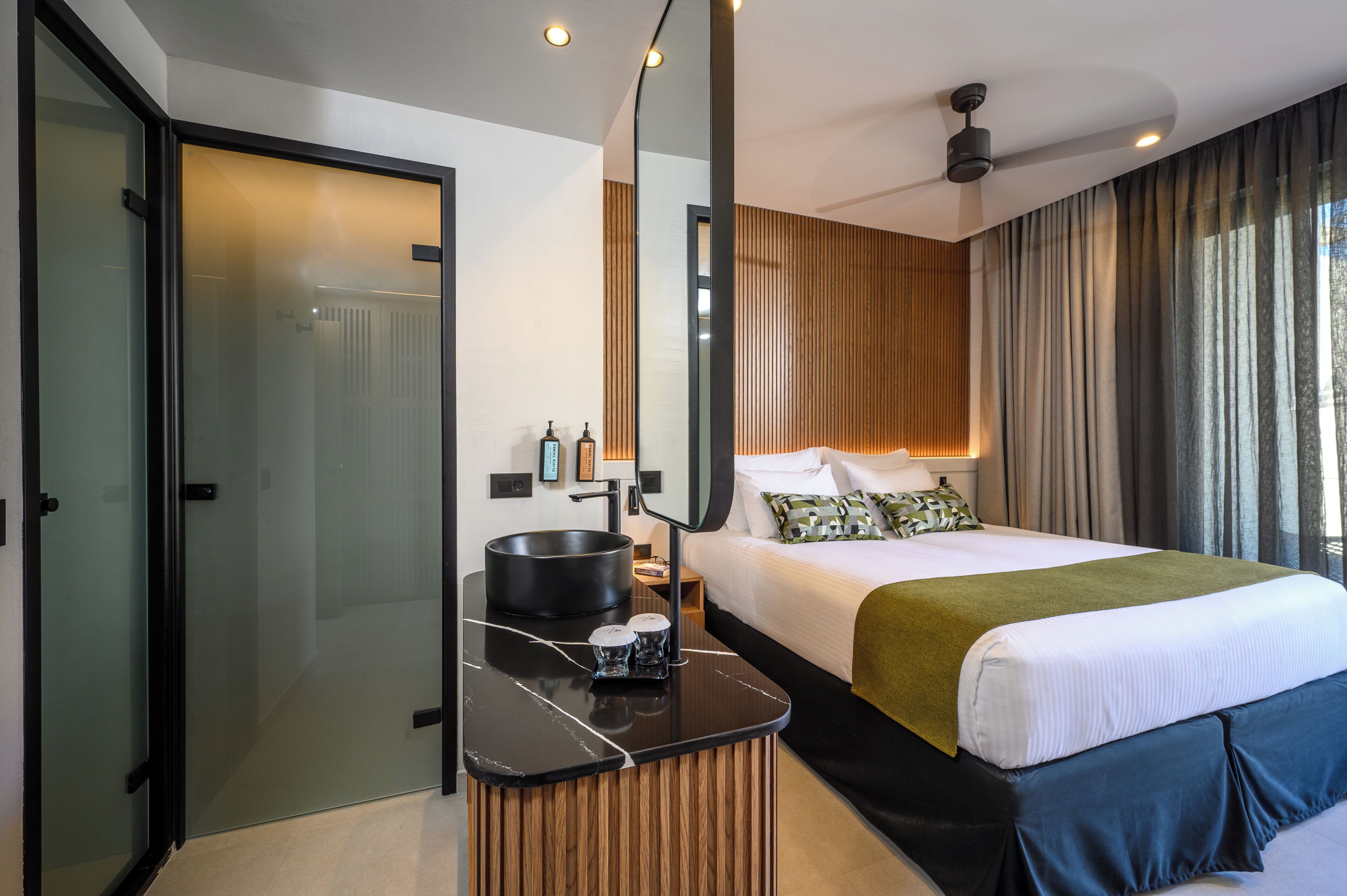 A modern hotel room with a bed, a bedside table with a mirror, a small sink, and a view of the outdoors.