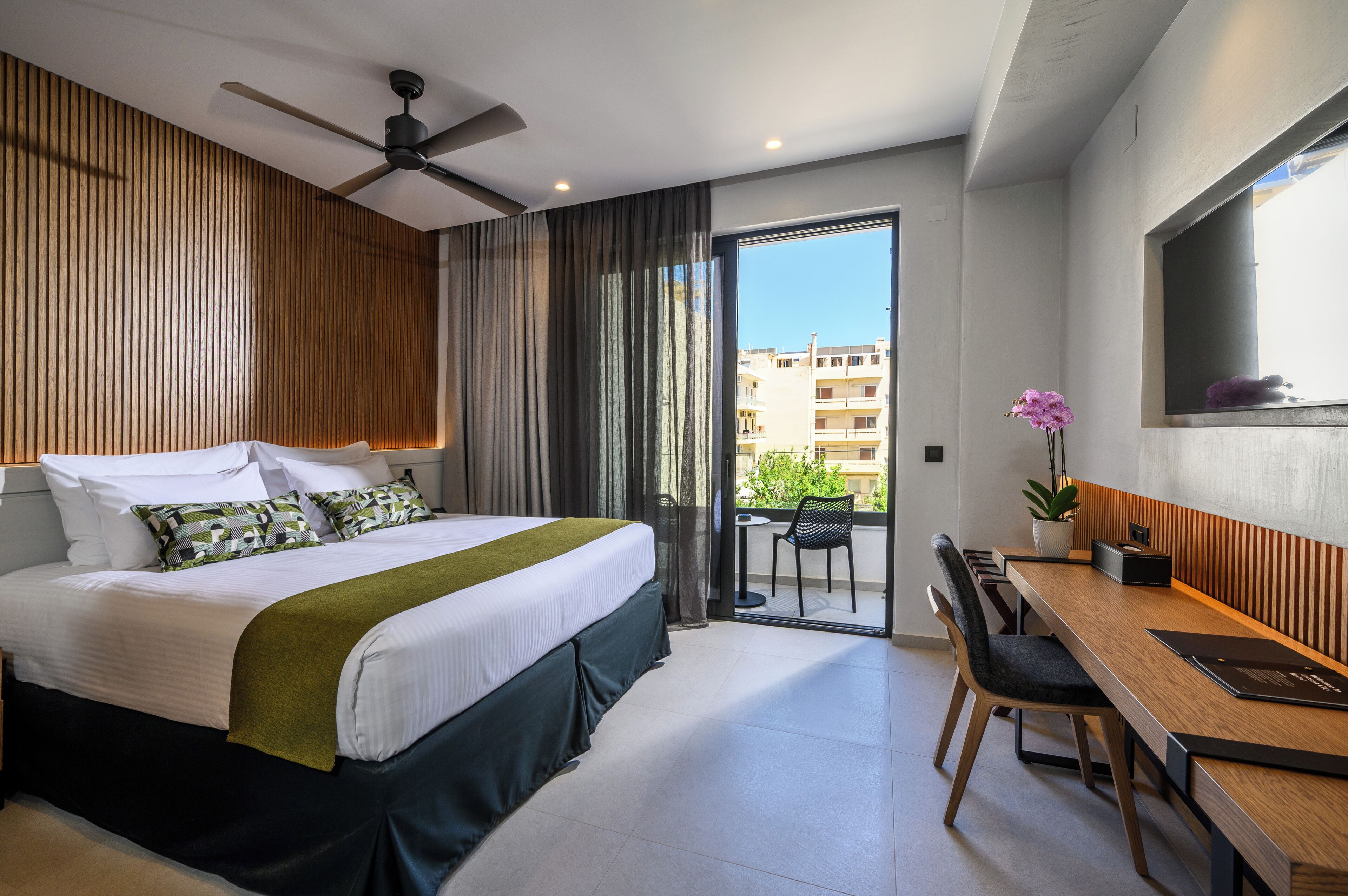 Quarto de hotel com cama, escrivaninha, cadeira, ventoinha no teto, varanda com vista para edifícios e planta em vaso.