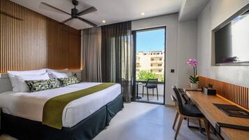 Quarto de hotel com cama, escrivaninha, cadeira, ventoinha no teto, varanda com vista para edifícios e planta em vaso.