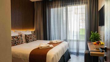 A modern hotel room with a large bed, a bedside table with a fruit bowl, and a view of the outdoors through sheer curtains.