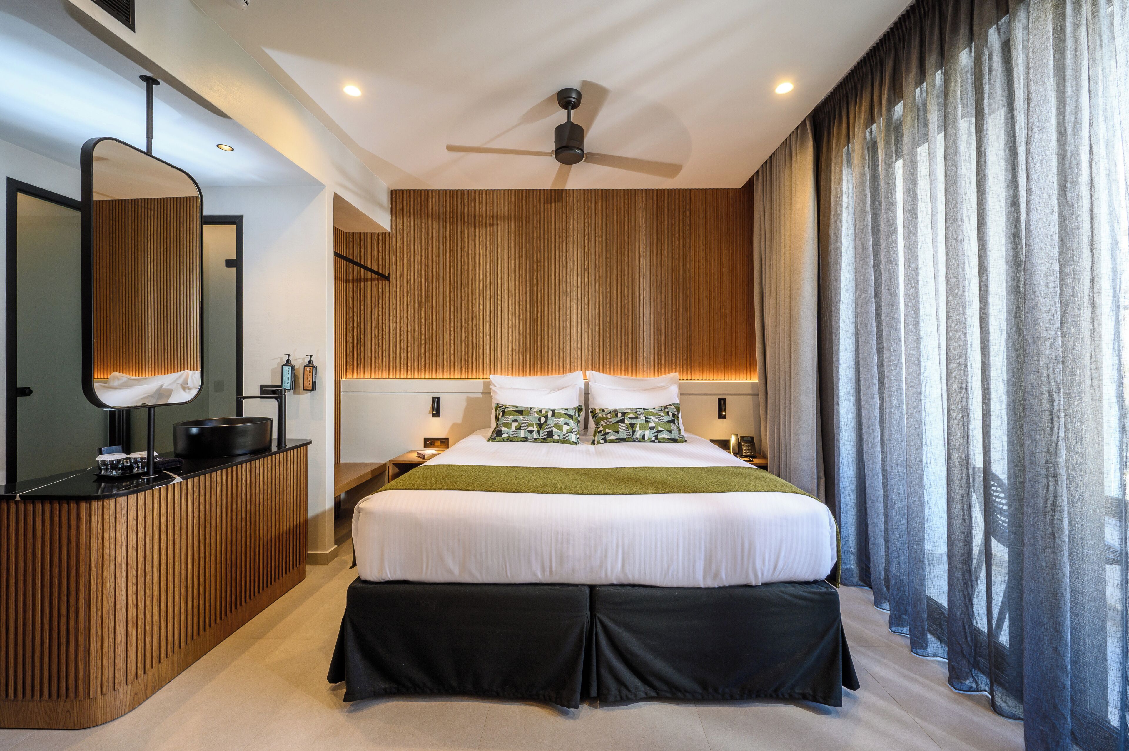 A modern bedroom with a large bed, a wooden desk, a ceiling fan, and a view of the outdoors through sheer curtains.