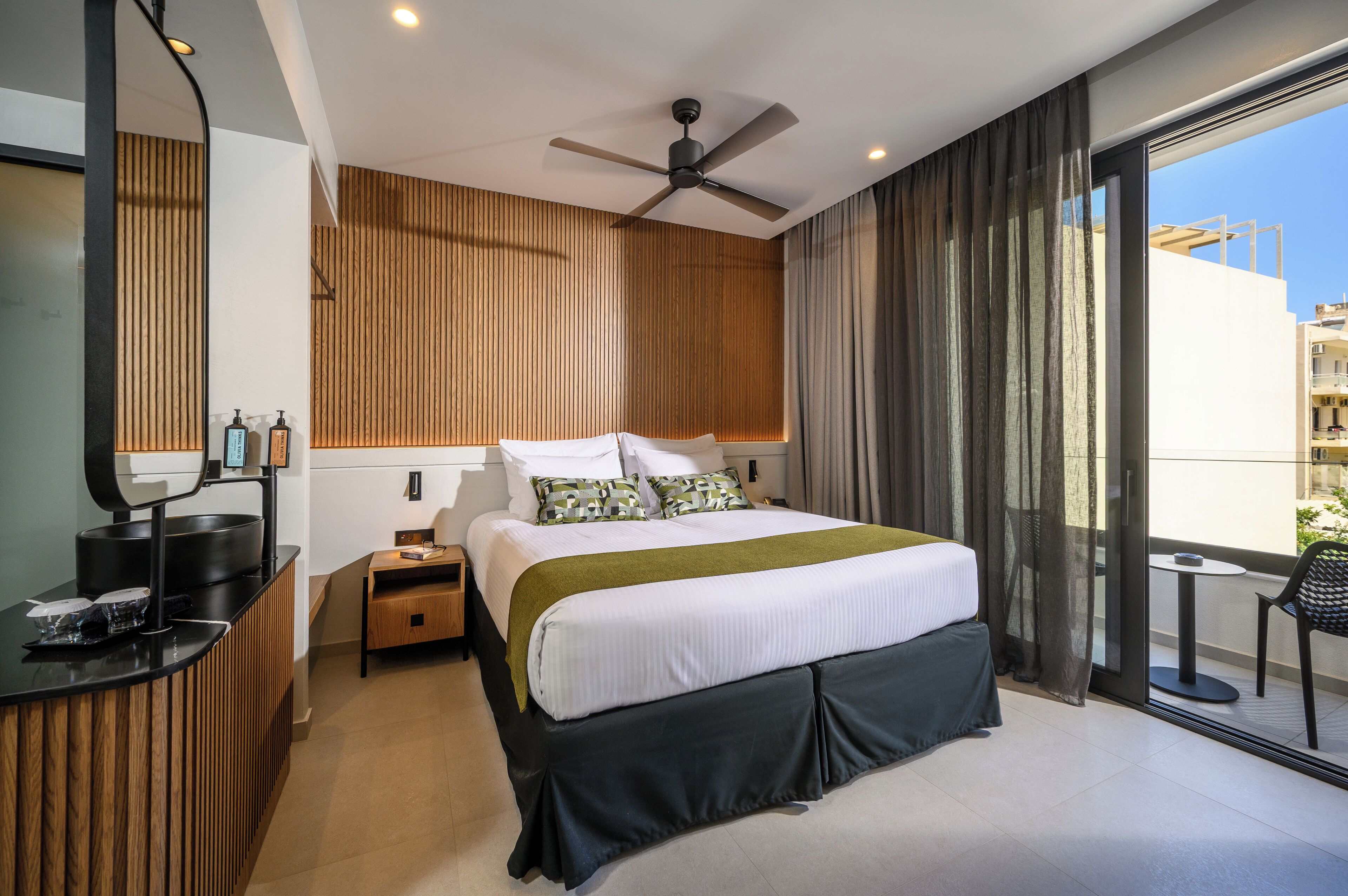 A modern hotel room with a bed, a desk, a mirror, a ceiling fan, and a view of the outside.