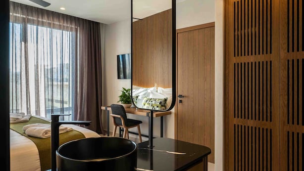 A modern hotel room with a bed, a desk with a chair, a large window with curtains, a wooden wardrobe, and a black basin on the counter. - Anesea City (Heraklion)
