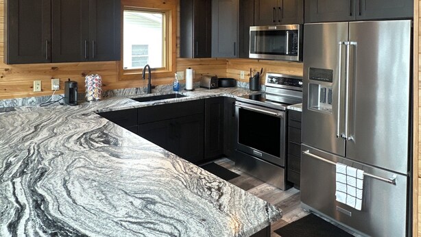 A modern kitchen with dark cabinets, stainless steel appliances, and a marble countertop. - Lakefront Escape with Loft & Summer Bunkhouse — Steps from the Water & Trails! (Waubun)
