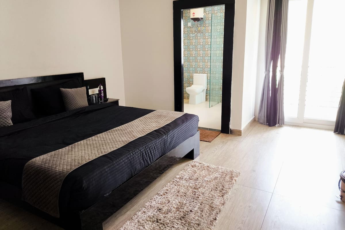 A bedroom with a large bed, a wooden headboard, a small bedside table, and a bathroom visible through a glass door.