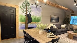 Een eetgedeelte met een houten tafel, zwarte stoelen, een beer klimt over een muurschildering van een boom, een grijze bank en een raam met uitzicht op bomen.
