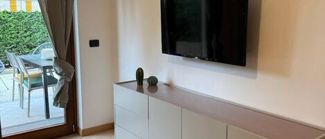 A modern living room with a flat-screen TV mounted on the wall, a white cabinet below, and a sliding glass door leading to an outdoor seating area.