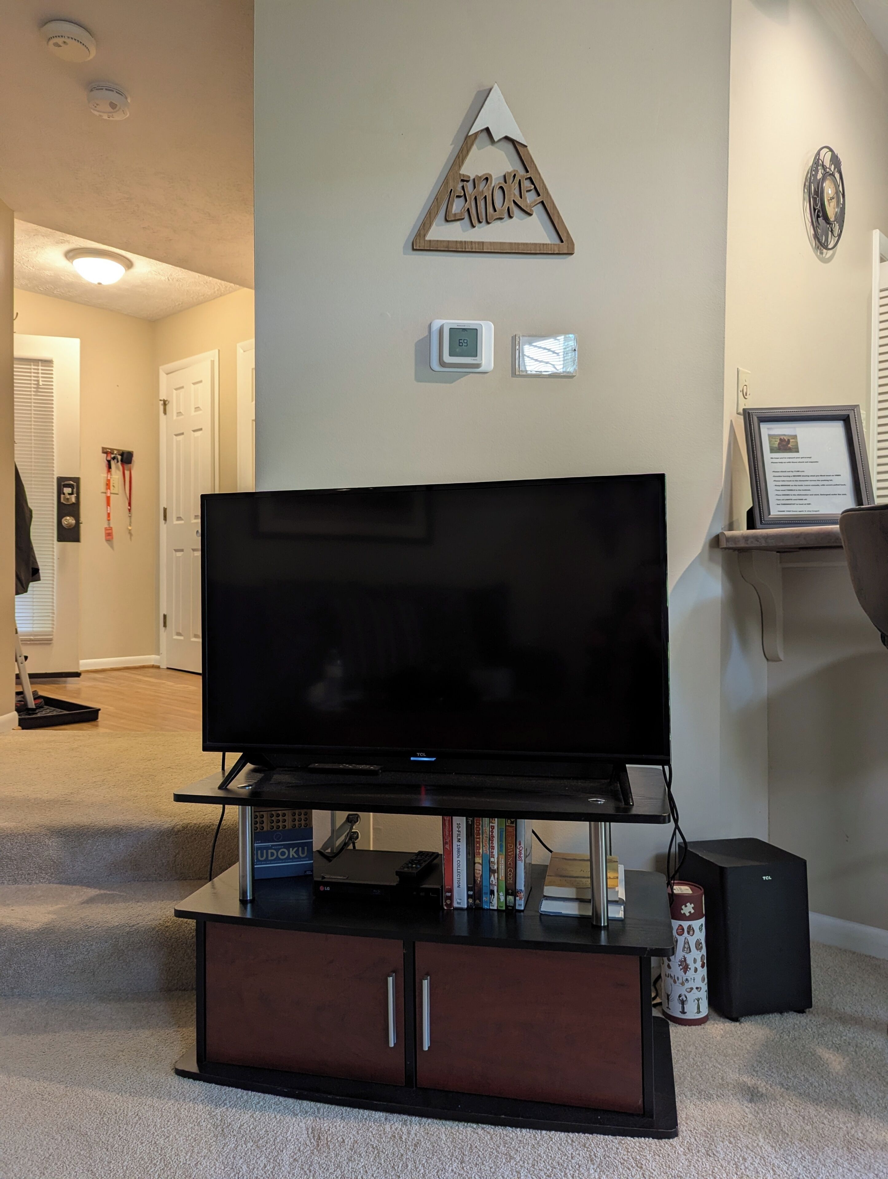 A living room with a flat-screen TV on a wooden stand, a small shelf with books, a framed certificate, and a wall-mounted thermostat.