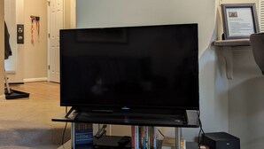 Ein Wohnzimmer mit Flachbildfernseher auf einem Holzständer, einer kleinen Vitrine mit Dekoration und einem an der Wand montierten Thermostat.