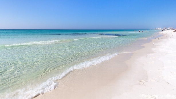 Une plage à l’eau turquoise et limpide, au sable blanc, et sous un ciel dégagé.