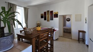 Un comedor con una mesa de madera, sillas, una planta en maceta, una jarra amarilla y una cesta. Se aprecia una ventana con cortinas, una lámpara y una obra de arte en la pared.