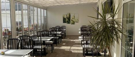 A spacious indoor area with large windows, multiple tables and chairs, and a potted plant.