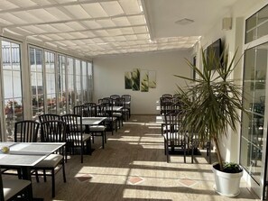 A spacious indoor area with large windows, multiple tables and chairs, and a potted plant.