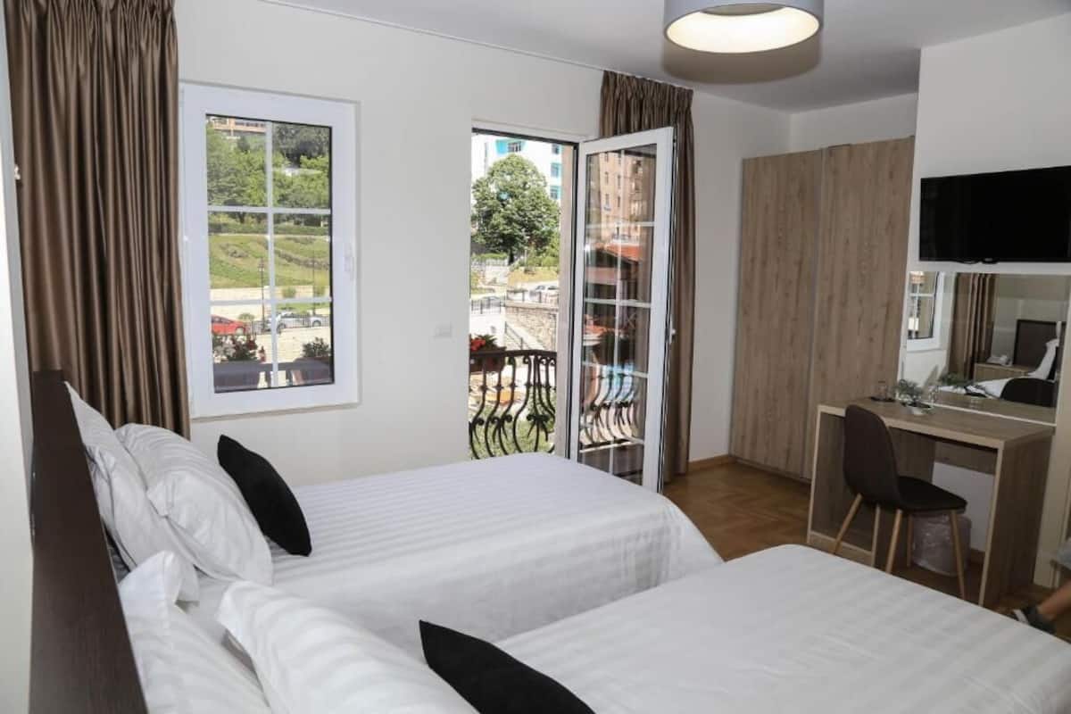A hotel room with two beds, a desk, a TV, and a balcony with a view of the city.
