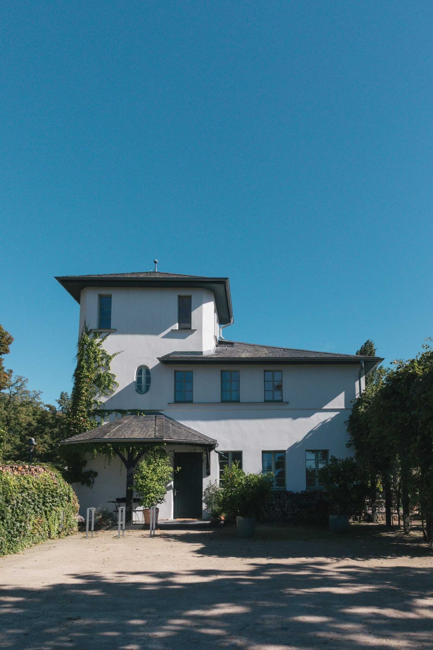Ein zweistöckiges Gebäude mit symmetrischer Fassade, einem überdachten Eingangsbereich und einem kleinen Garten mit Topfpflanzen.