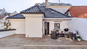 En täckt uteplats med ett trädäck, en grill, utemöbler och en krukväxt.