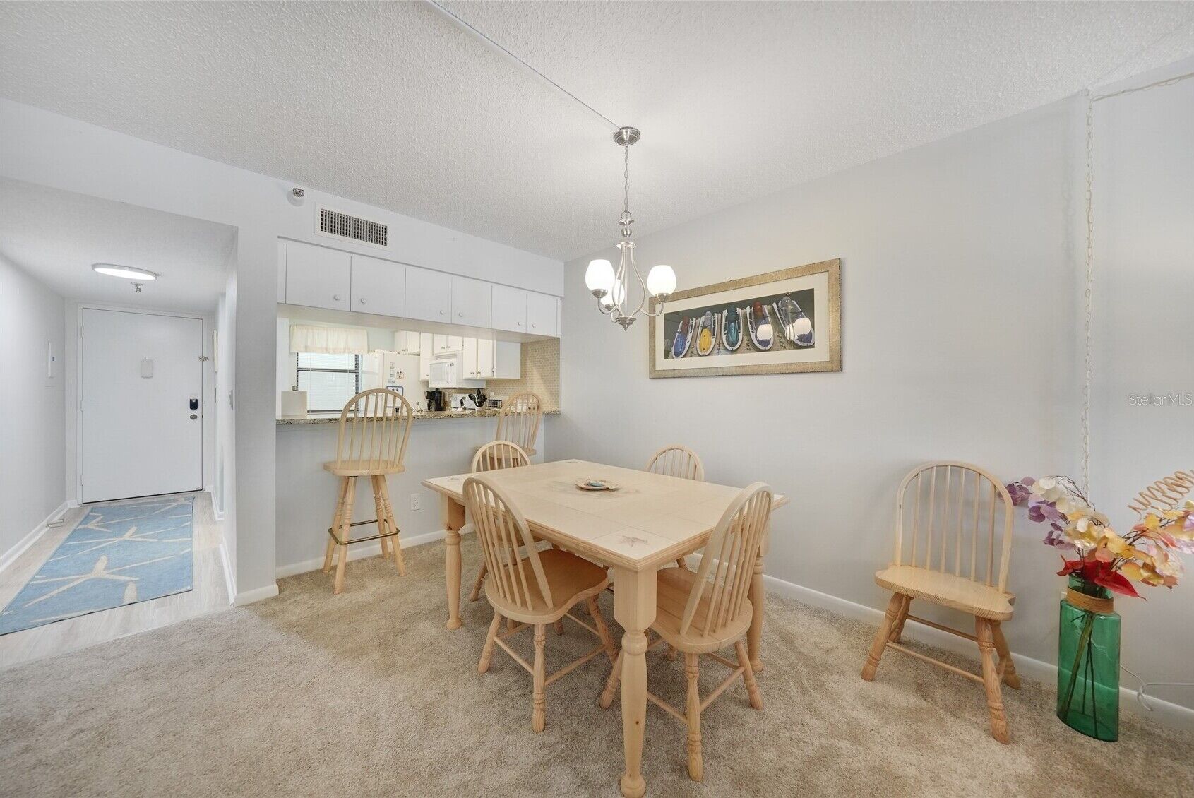 A dining area with a wooden table and chairs, a kitchen in the background, and a framed artwork on the wall.