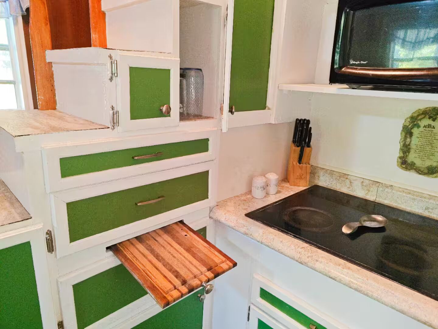 Une cuisine avec des meubles de rangement verts et blancs, un four à micro-ondes et un tiroir contenant une planche à découper en bois.