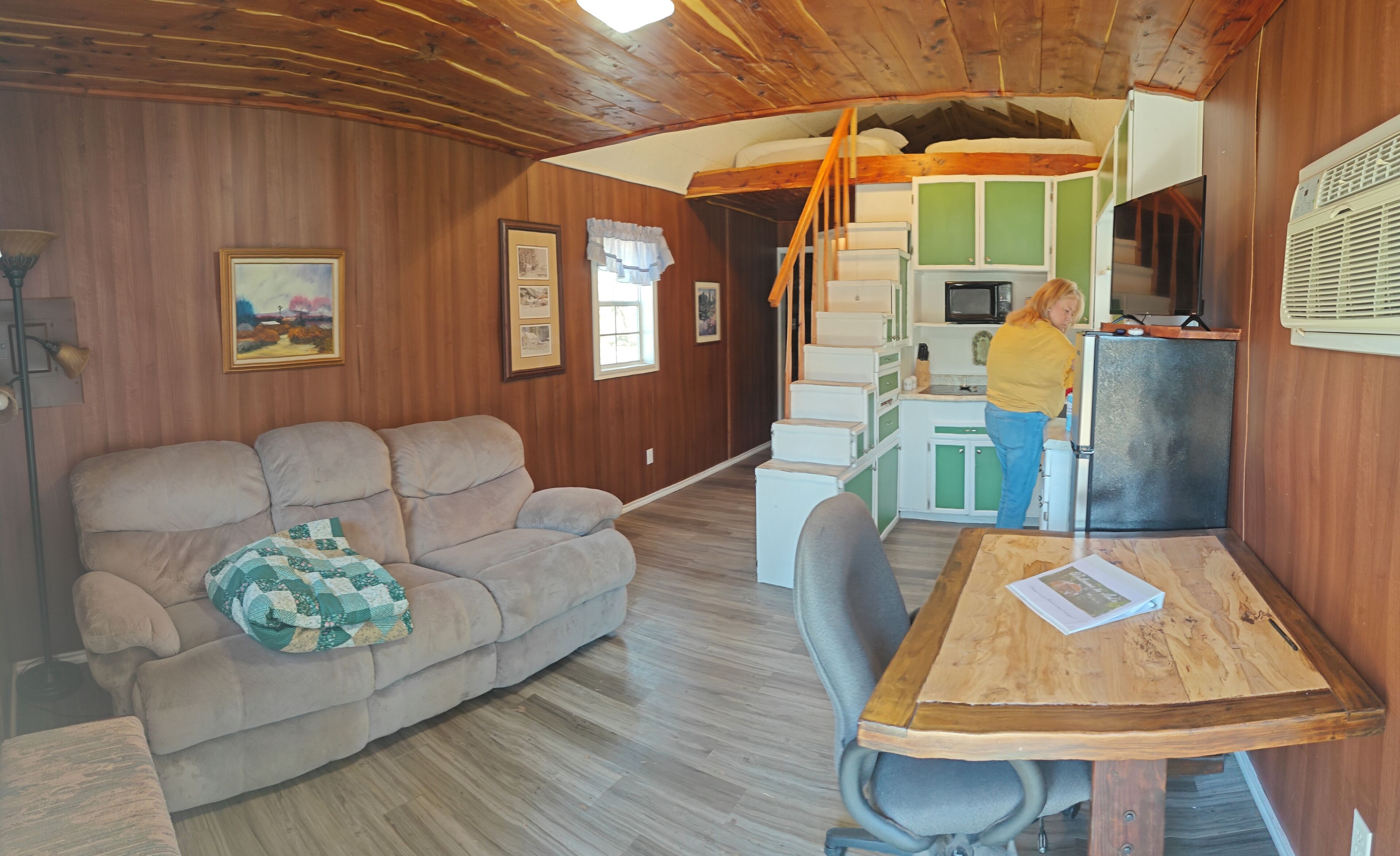 A cozy cabin interior with a wooden ceiling, a sofa, a dining table, and a kitchen area.