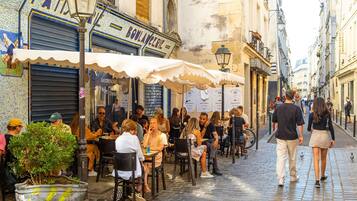 A bustling outdoor café with patrons seated at tables, surrounded by buildings with various signs and a cobblestone street.
