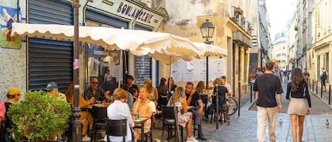 Un café animé en plein air, avec des clients assis à des tables, entouré d'immeubles ornés de diverses enseignes et bordé d'une rue pavée.