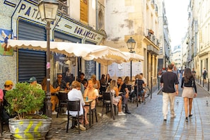 A bustling outdoor café with patrons seated at tables, surrounded by buildings with various signs and a cobblestone street.