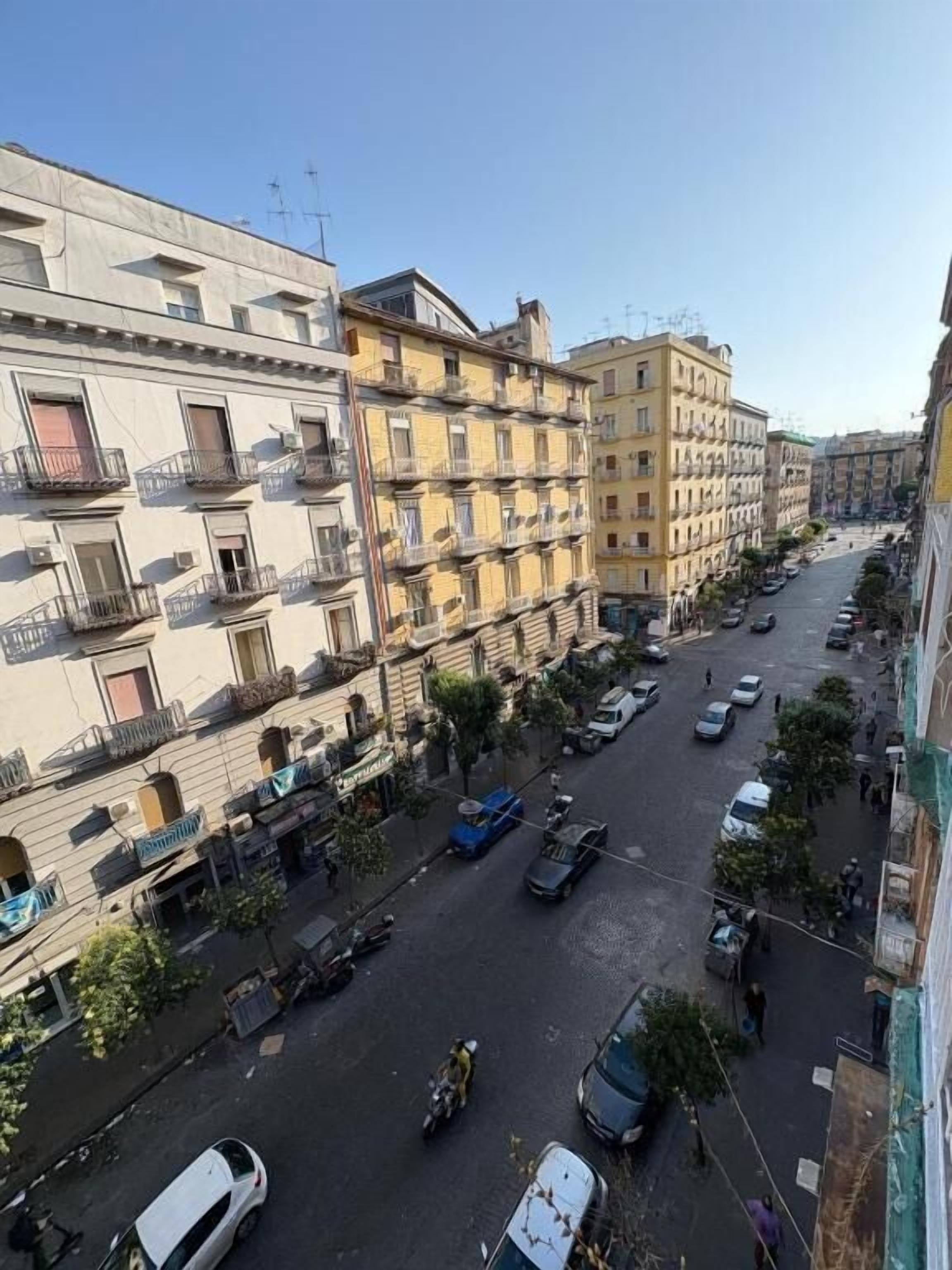 A multi-story residential building with balconies, a street with parked cars, and a clear sky.