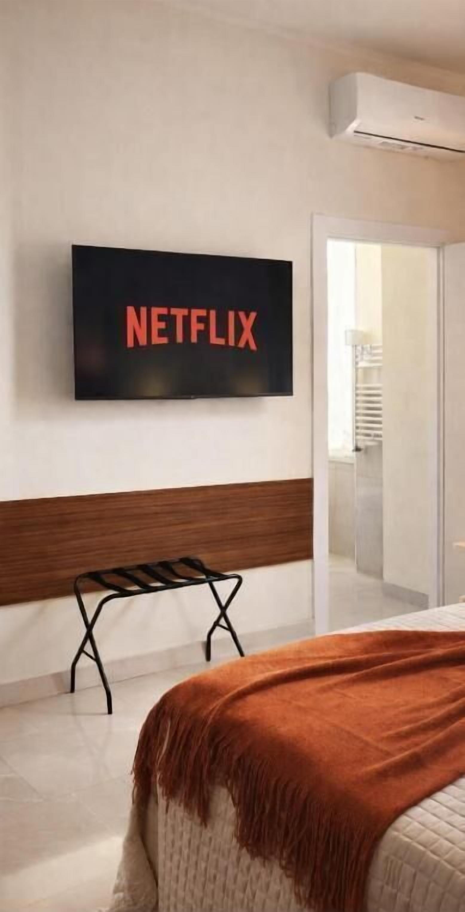 A bedroom with a bed, a bench, a wall-mounted TV displaying 'NETFLIX,' and an air conditioning unit.