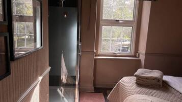 Ein Zimmer mit Dusche, einem Bett mit gesteppter Decke und einem Fenster mit blickdichtem Vorhang.